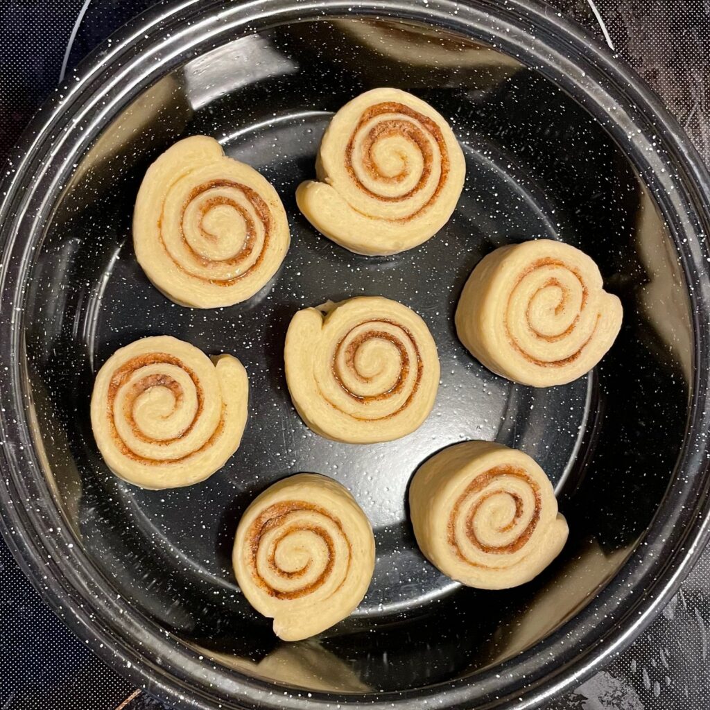 Seven cut cinnamon roll spirals arranged in a round pan, unbaked, the cinnamon swirl visible