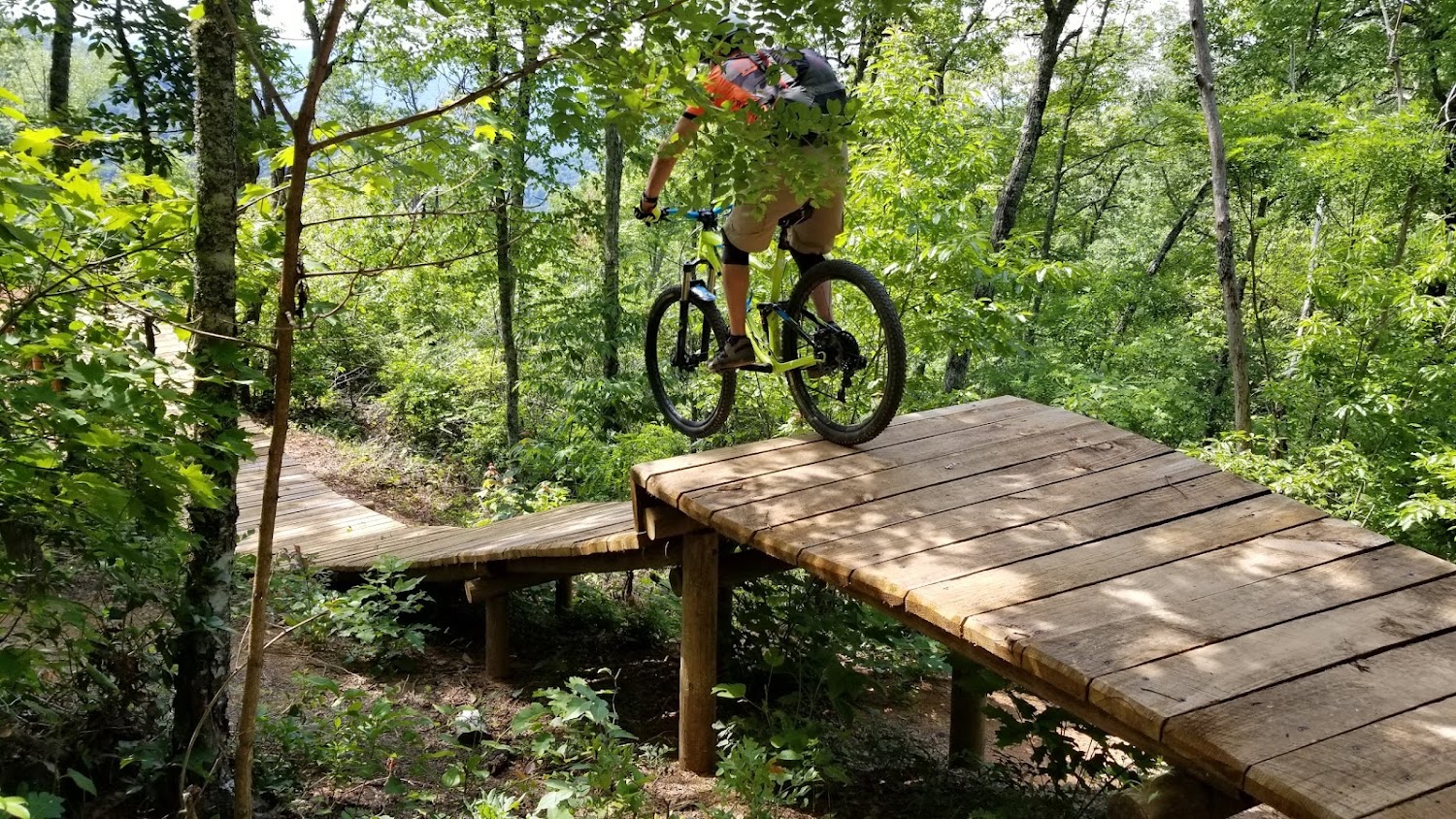 A rider crosses a raised wooden boardwalk feature on a green, wooded trail at Fire Mountain.