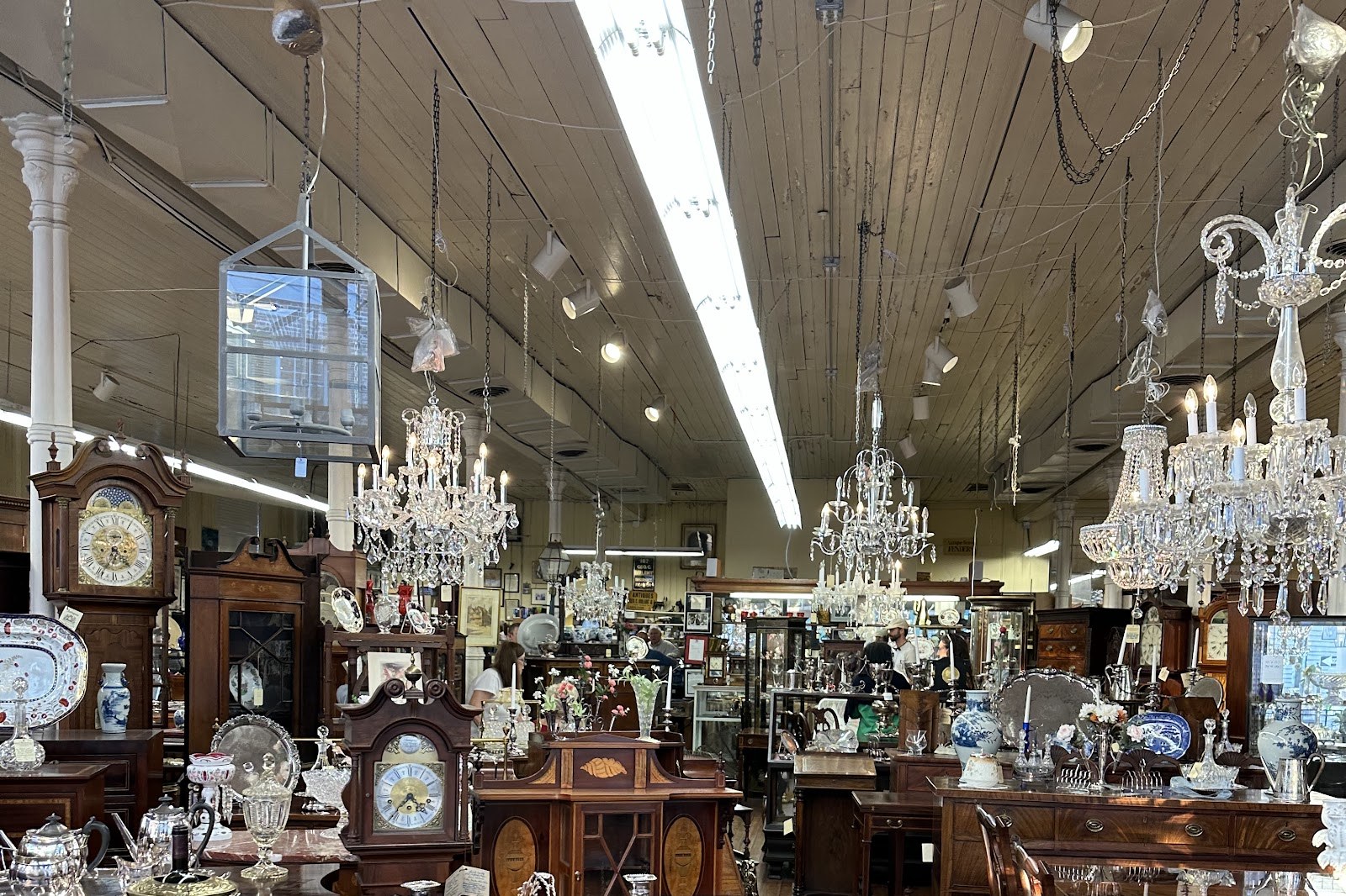 Crystal chandeliers hanging in rows under a plank ceiling at George C. Birlant and Co, with longcase clocks, mahogany chests and silver on the tables below.