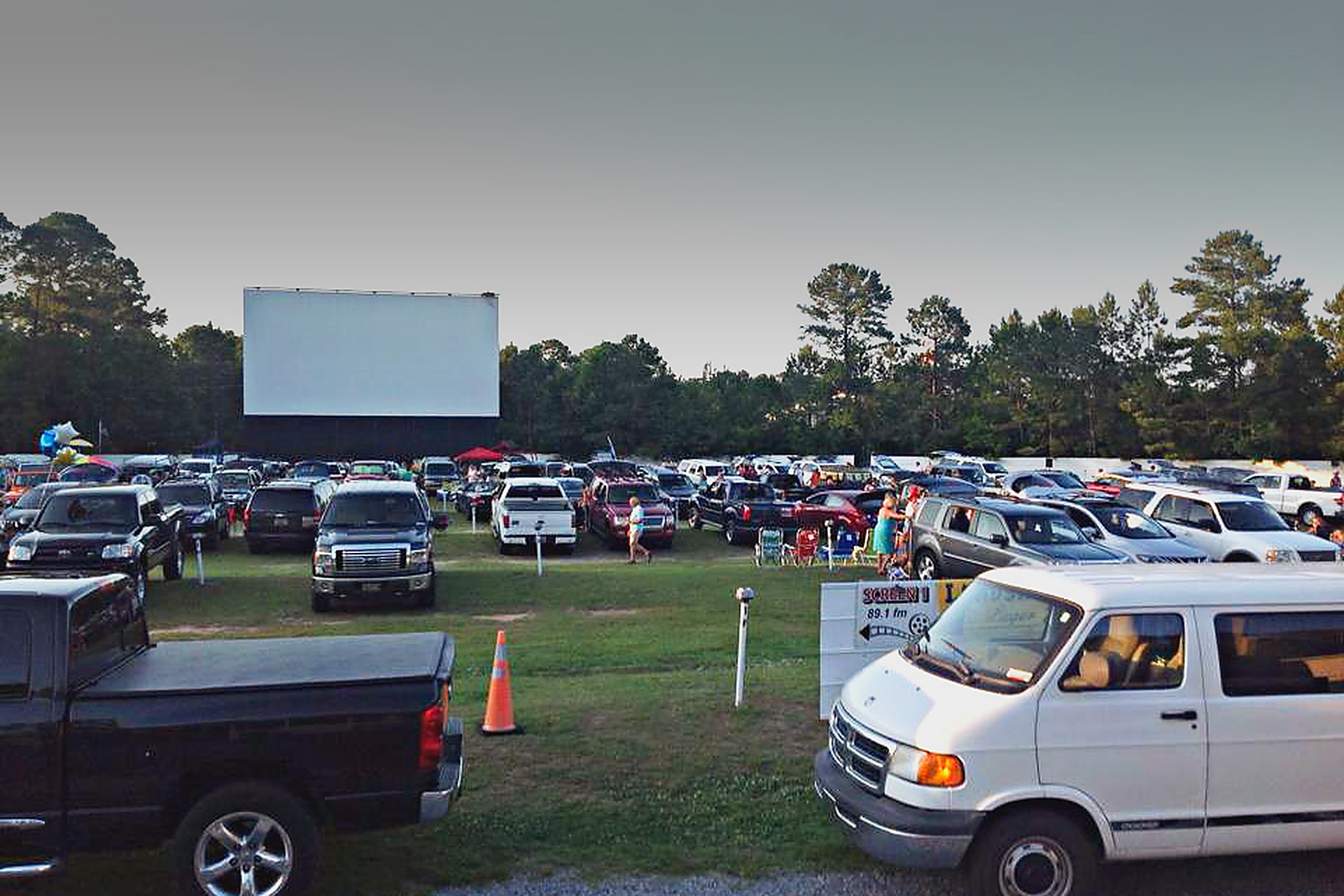 Cars parked in rows on the grass at the Highway 21 Drive-In in Beaufort, the screen standing against pine trees while families set out chairs before the film.