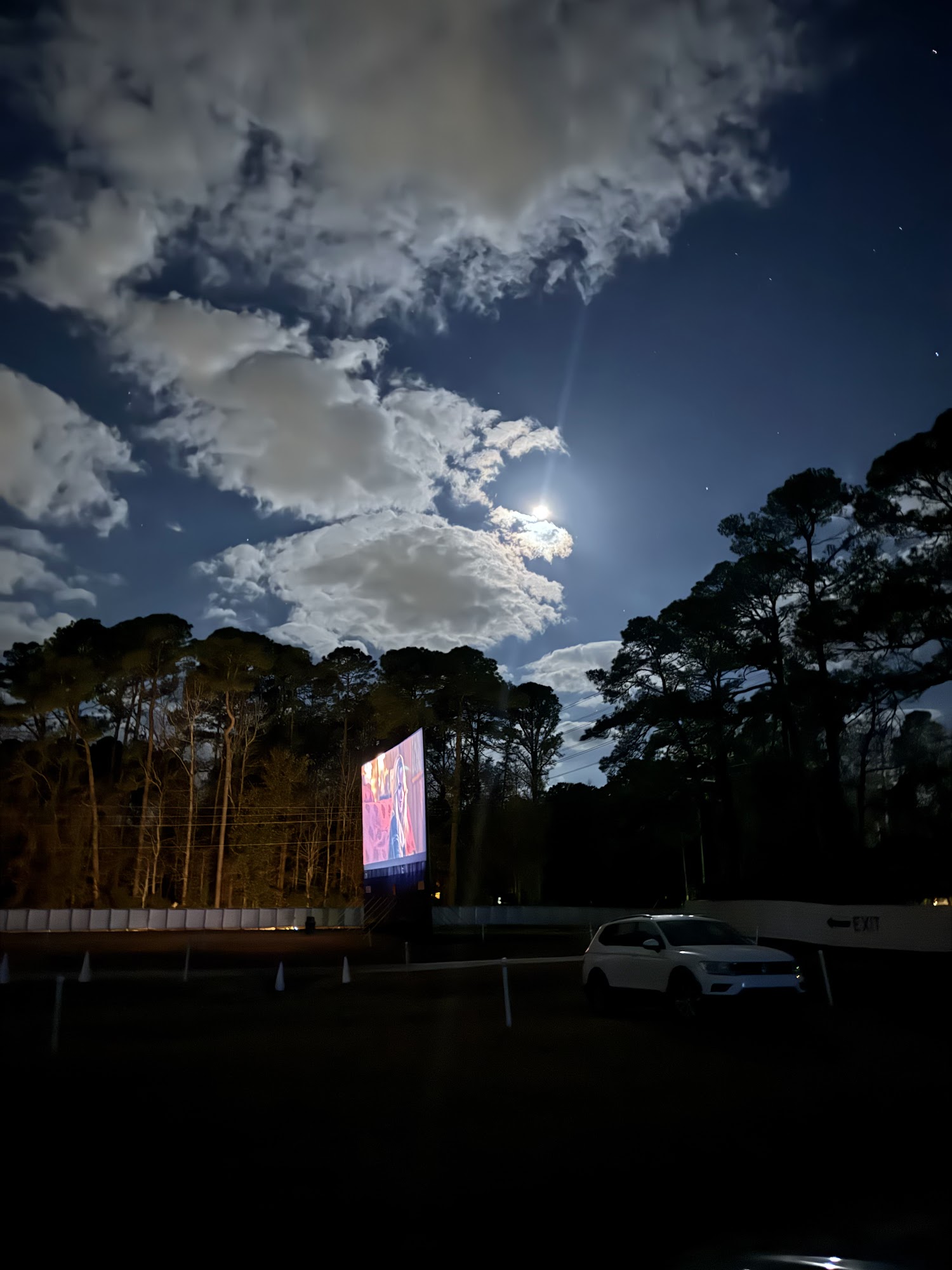 The lit screen of the Highway 21 Drive-In at night, a full moon breaking through cloud above the pines and one car parked on the field.