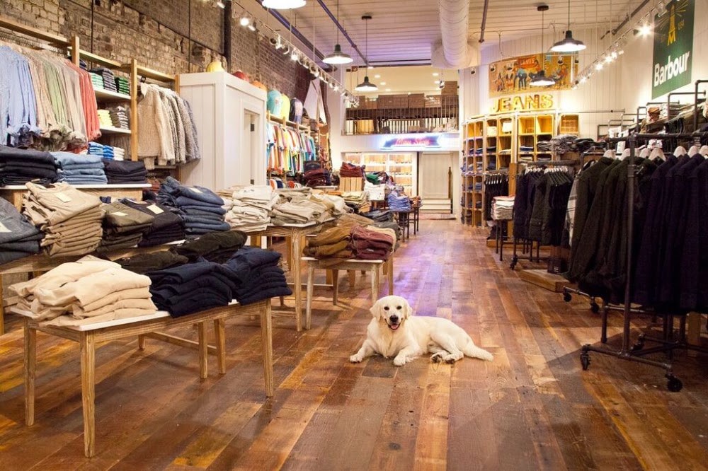 Folded knitwear stacked on wooden tables at M. Dumas and Sons, rails of coats along a brick wall, and a golden retriever lying on the plank floor.