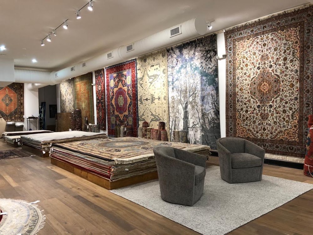 Hand-knotted rugs hung floor to ceiling along a white wall at Michael Mitchell, with stacks of rugs on a low plinth and two grey swivel chairs.