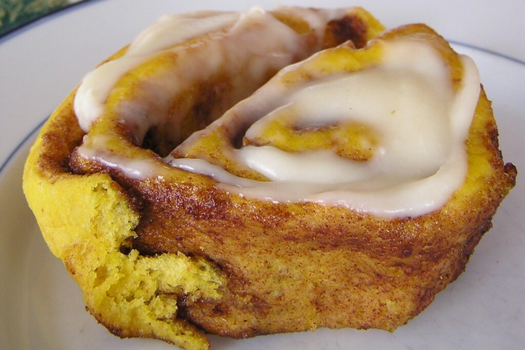 A single pumpkin cinnamon roll on a white plate, cream cheese icing pooled over the coils