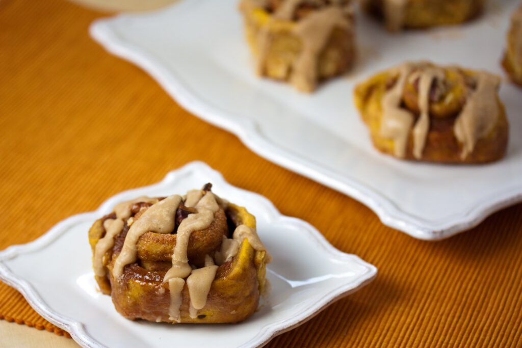 A pumpkin cinnamon roll on a white plate with more rolls on a platter behind it