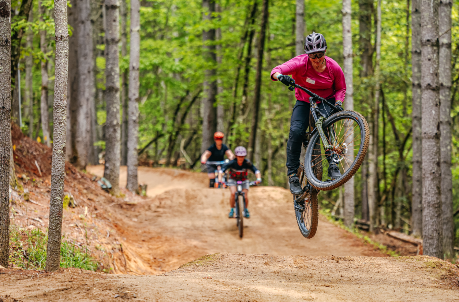 Three riders descend a bermed dirt trail through tall pines at Ride Kanuga, the closest rider lifting the front wheel.