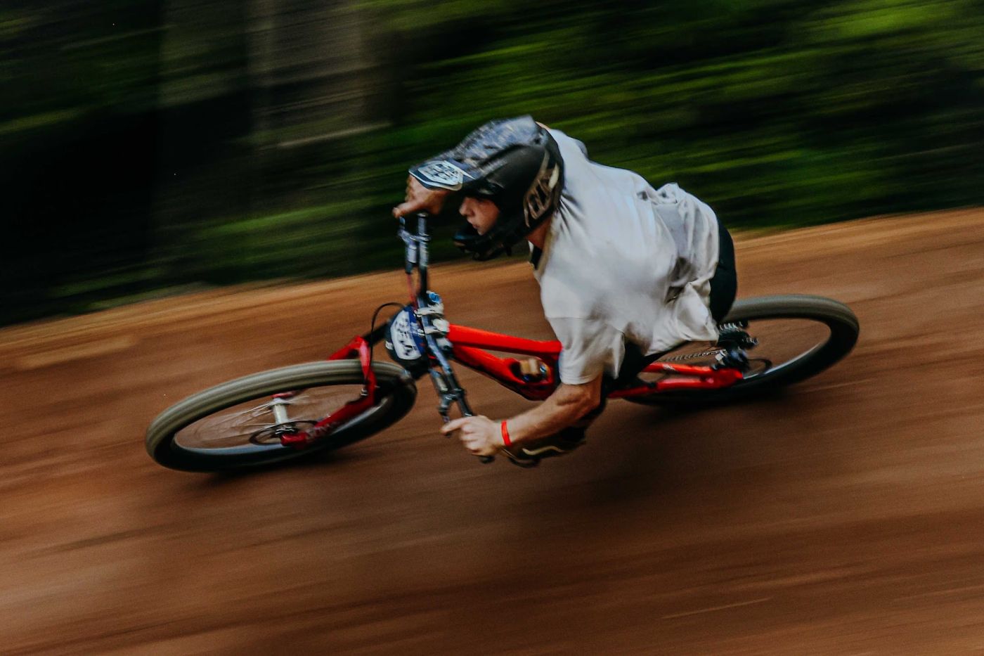 A mountain biker leans hard through a banked dirt turn on a downhill trail, the forest blurred behind them.