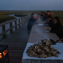 A shovel tipping steaming oysters onto a long communal table on a marsh dock at dusk, with a wood fire burning alongside.