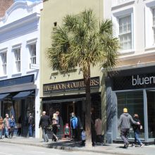 Shoppers walking the King Street pavement in Charleston past awnings and a palmetto, with the Half-Moon Outfitters and bluemercury shopfronts behind them.
